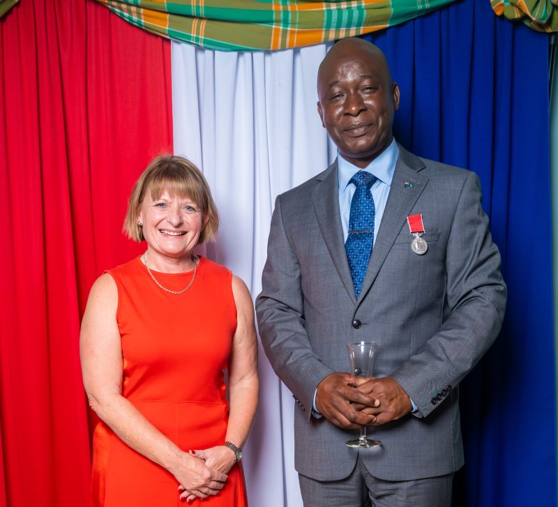 Lt Col Alvin Ryan BEM is wearing a gray suit with the new medal and Governor Sarah Tucker wears a red sleeveless dress.