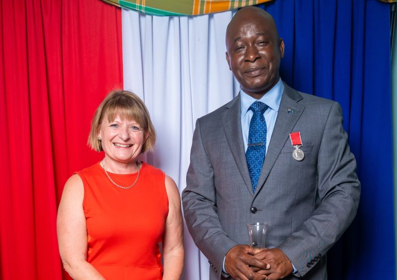 Lt Col Alvin Ryan BEM is wearing a gray suit with the new medal and Governor Sarah Tucker wears a red sleeveless dress.